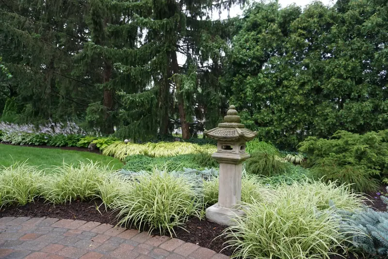 A curved brick paver walk beside ornamental grasses, a stone lantern and tall evergreens
