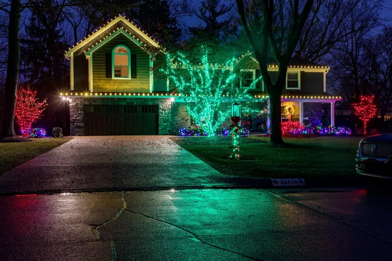 A house at night lit by English Gardens: warm white rooflines, a tree wrapped in green lights and red-lit shrubs